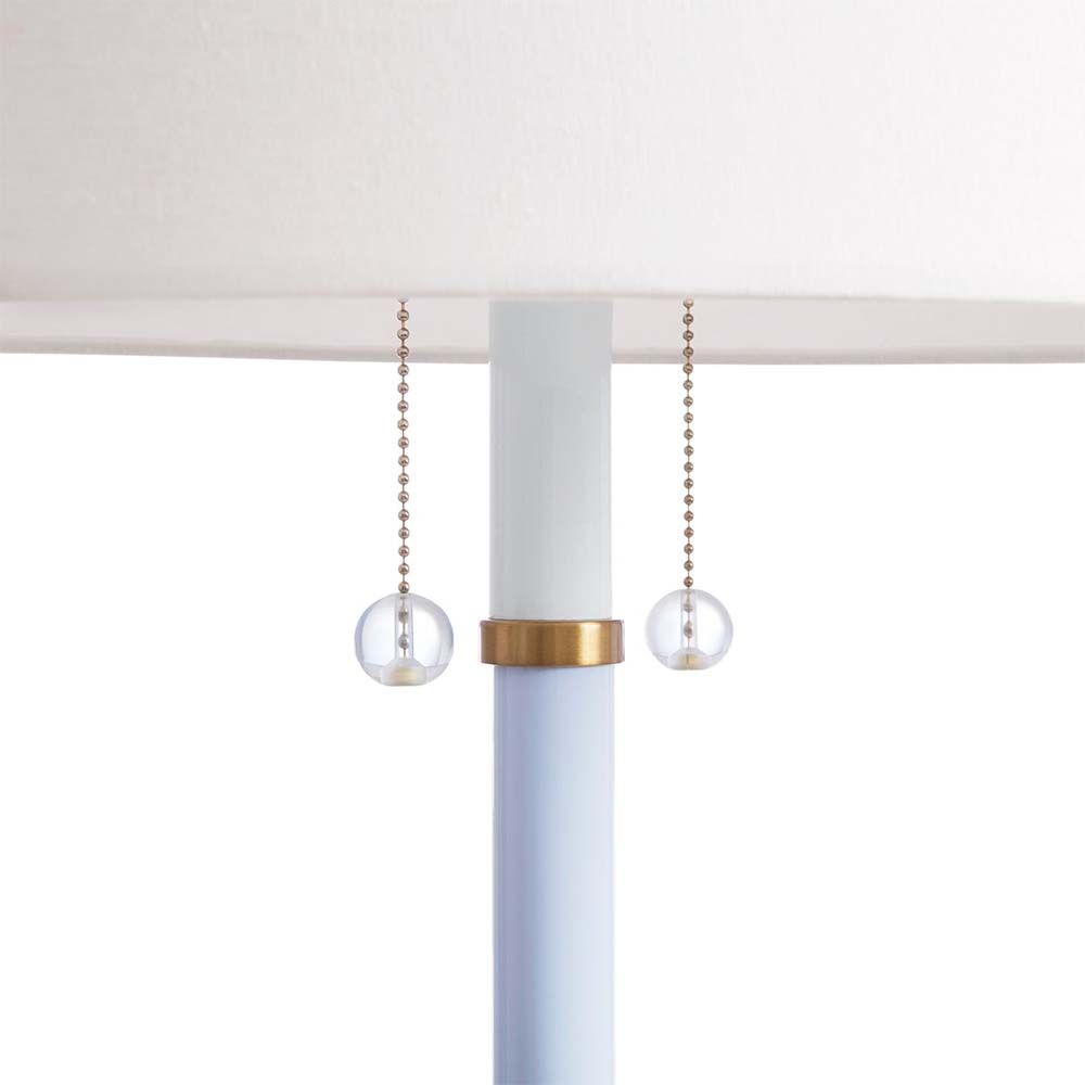 A blue-hued three footed floor lamp with pull strings, brass elements and a white shade.