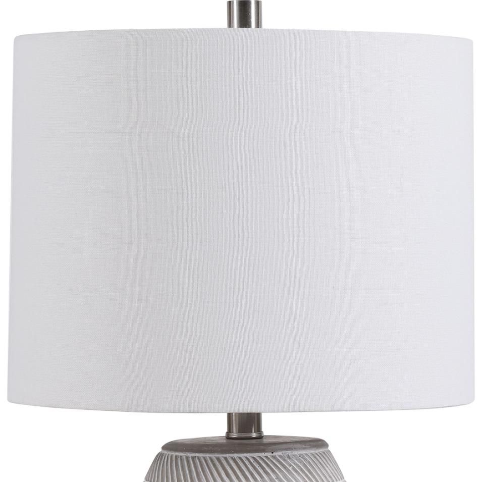 This table lamp has a concrete base with white etching, brushed nickel accents, and a light beige linen shade, offering modern sophistication and understated elegance.